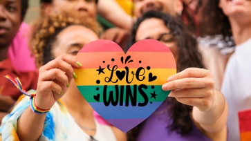 Love Wins Pride Month image of two women holding a paper rainbow heart, standing in a crowd